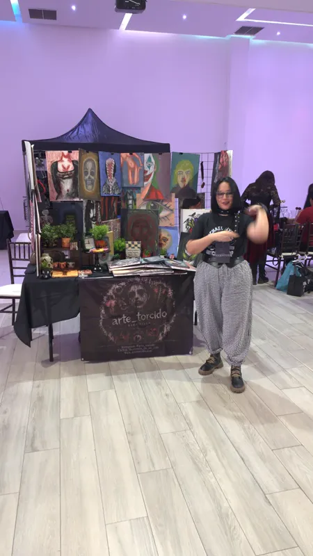 Sara Villa de pie frente a su stand con carpa en la Expo Gótica, con la lona de arte_torcido en la mesa y las pinturas colgadas en la rejilla