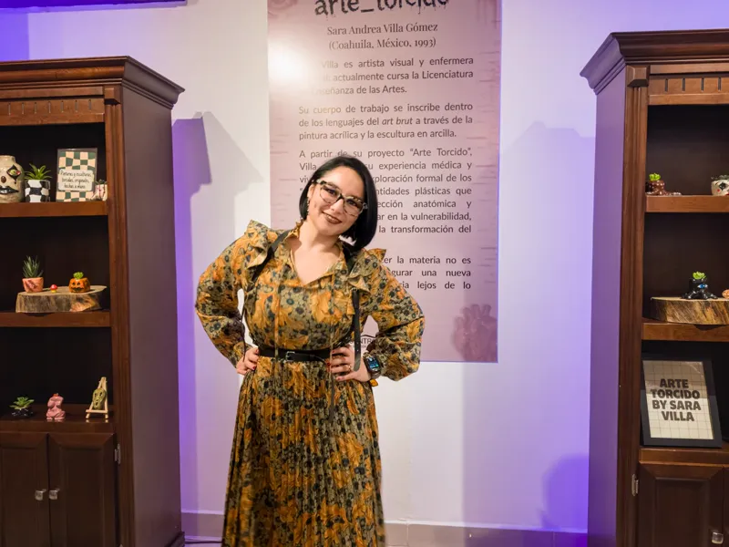 Sara Villa en la sala de exposición del CCLEMS, entre dos vitrinas de madera con seres torcidos de arcilla y frente al panel con su semblanza