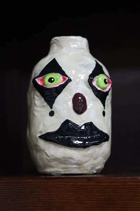 Ser torcido de arcilla: frasco con cara de payaso, ojos verdes y boca negra, en una vitrina de madera