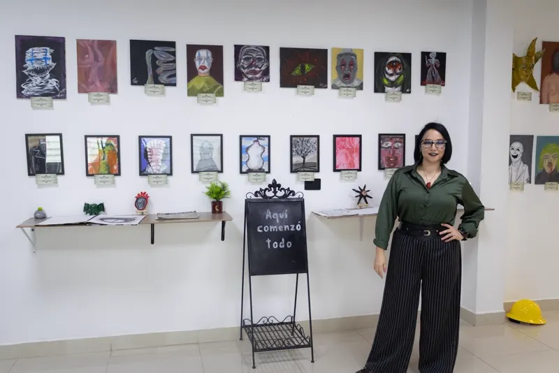 Sara Villa junto al muro blanco con dos filas de pinturas y un pizarrón que dice Aquí comenzó todo