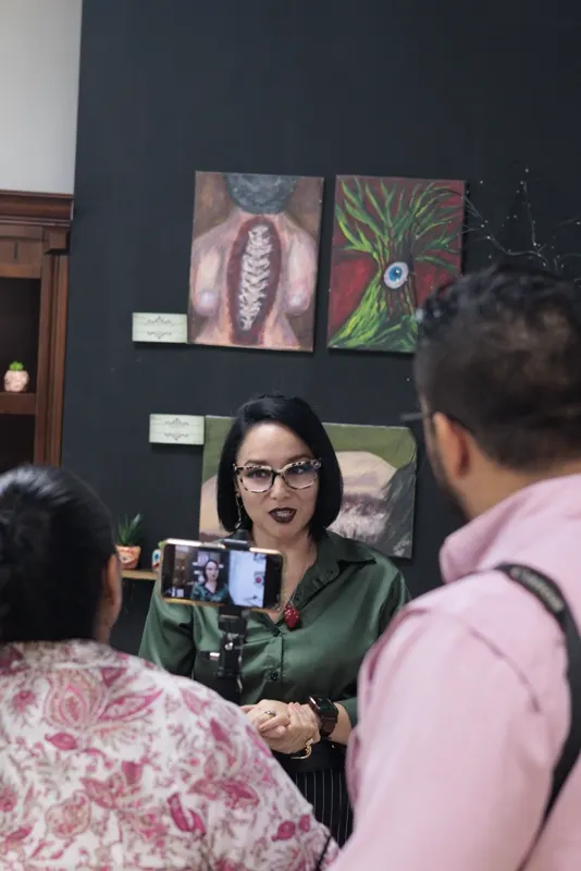 Un teléfono en tripié graba a Sara Villa mientras habla; detrás, sus pinturas en el muro negro