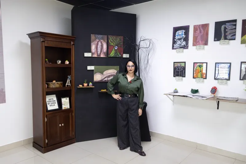 Sara Villa junto a la vitrina con el letrero Arte Torcido by Sara Villa y el muro negro con la espalda de columna expuesta y el árbol con un ojo
