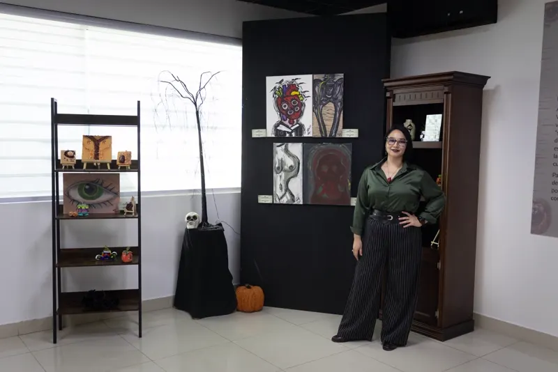 Sara Villa junto al muro negro con cuatro pinturas, entre ellas el rostro-corazón de tres ojos; a la izquierda, un estante con piezas de arcilla y un árbol negro