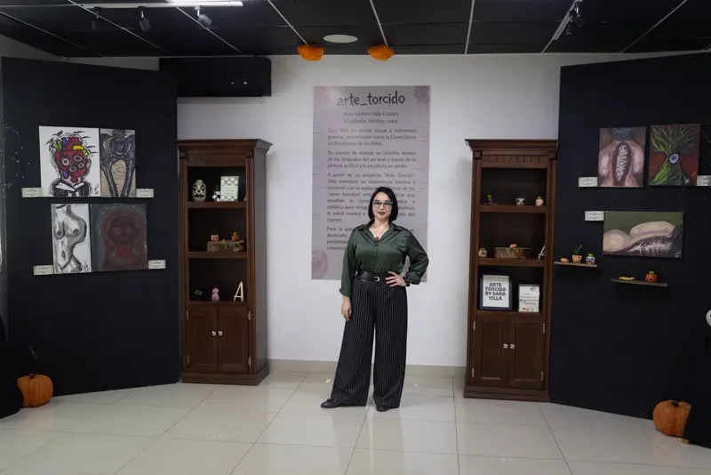 Sara Villa de pie al centro de la sala, entre dos vitrinas de madera con piezas de arcilla y dos muros negros con pinturas; al fondo, el panel arte_torcido con su semblanza