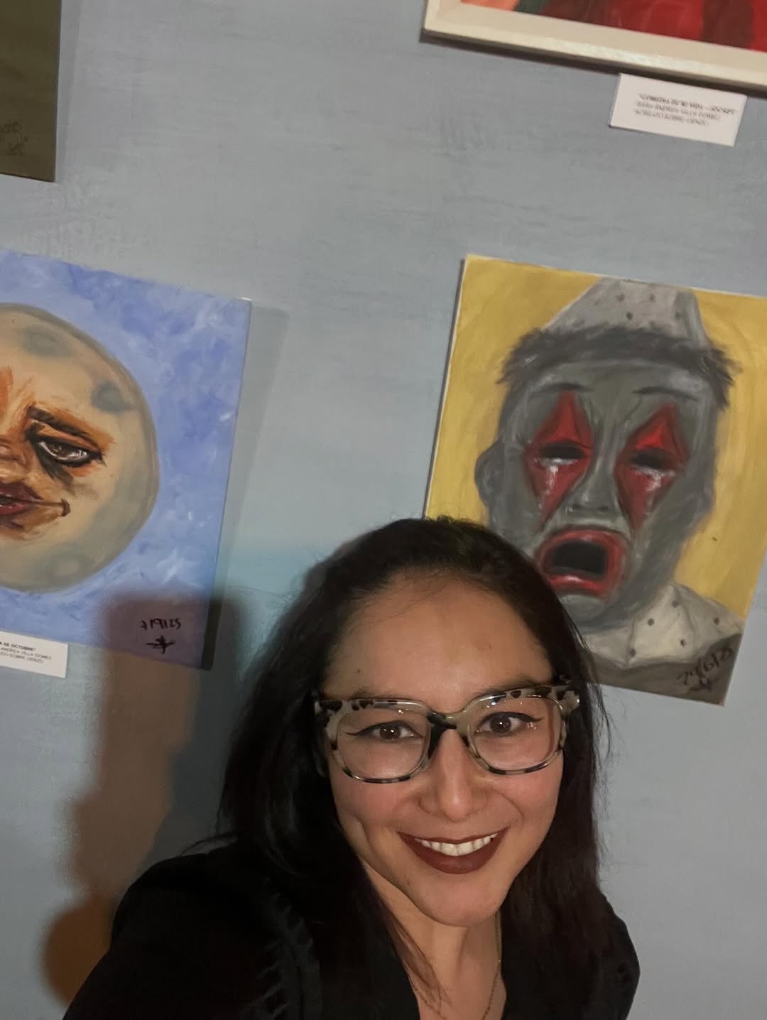 Sara Villa sonríe frente a sus pinturas de una luna con cara y un payaso